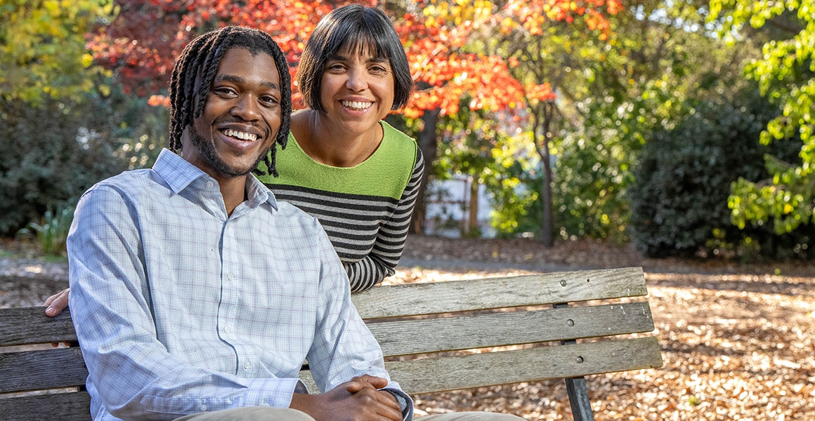 man and a woman outside on a bench smile at the camera